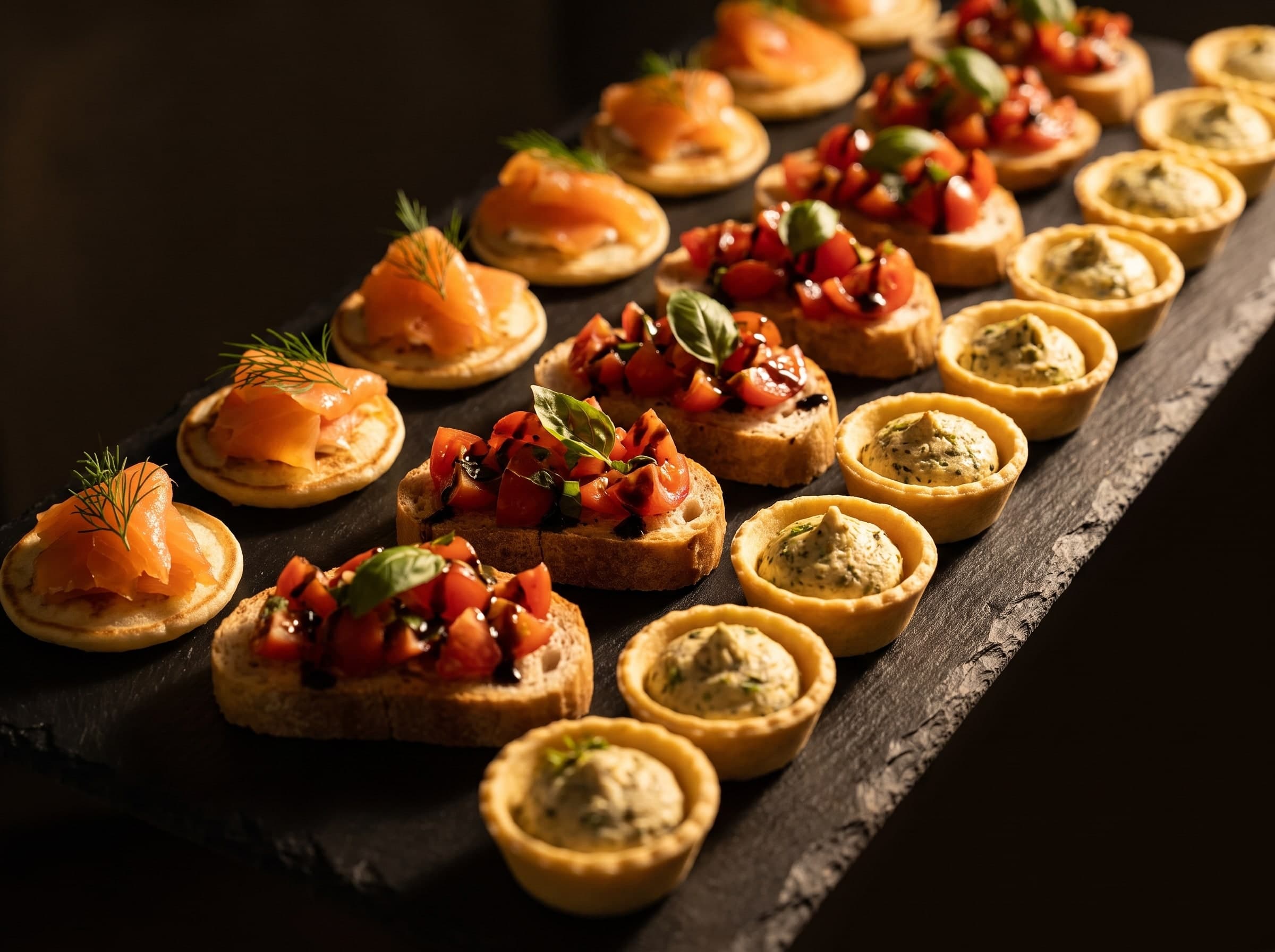 Planche d’ardoise sombre avec des rangées de blinis au saumon, de bruschettas et de tartelettes aux herbes