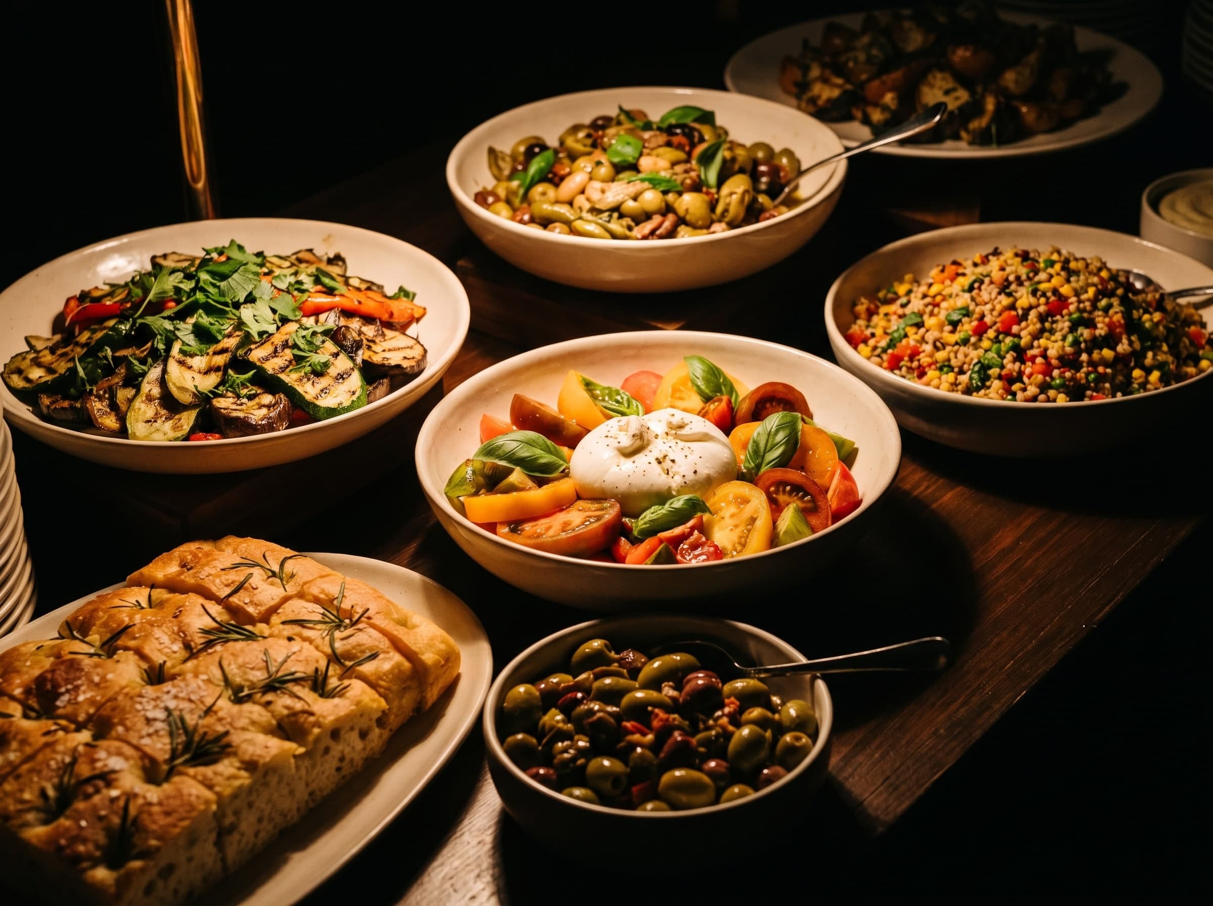 Buffet avec des plats de légumes grillés, une salade de burrata, une focaccia au romarin, des olives et une salade de céréales